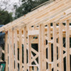 The photo shows the wood framing of a house under construction. A man is on a ladder at work on the home.