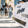 In the foreground is a close up of a yard sign that reads "Aquí Vote Here". In the background are students lined up outside to vote