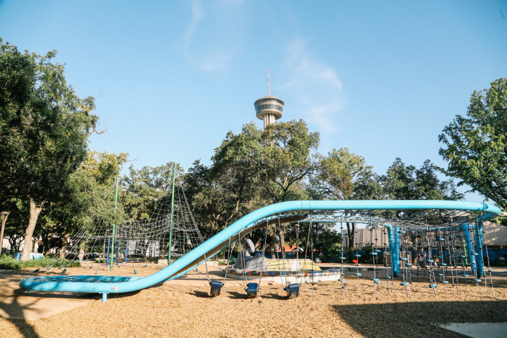 A wide shot of the play area, with swings, climbing ropes, climbing net, and more at Yanaguana Garden. The Tower of the Americas is seen in the background