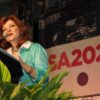 Carmen Tafolla reads her poem at a podium. In the background is an "SA2020" banner with colored icons representing the Cause Areas