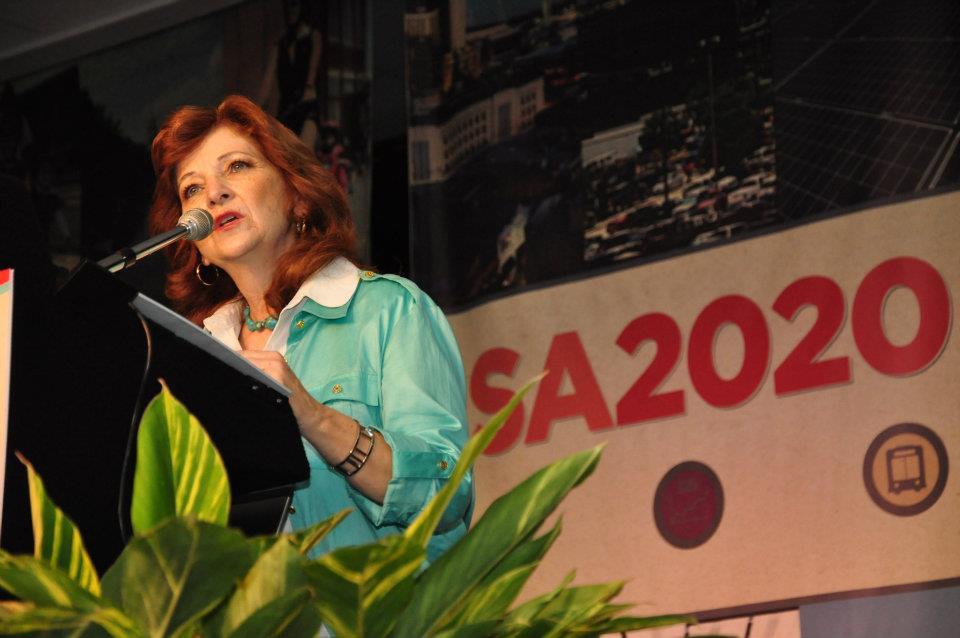 Carmen Tafolla reads her poem at a podium. In the background is an "SA2020" banner with colored icons representing the Cause Areas