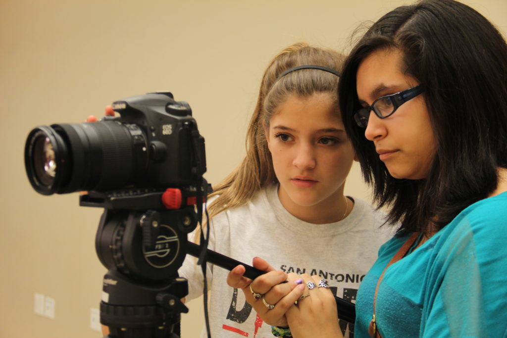 Two students stand behind a video camera, looking carefully at the shot